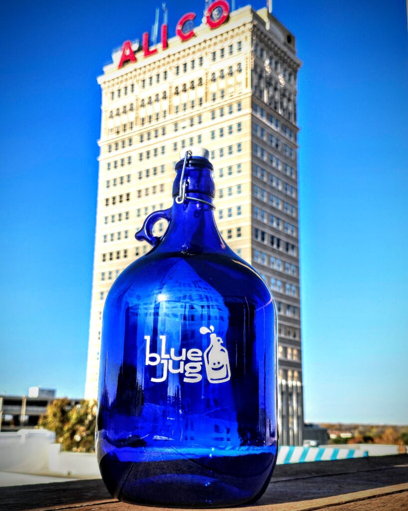 Blue glass water jug from Blue Jug of Waco positioned on a bridge walkway with a white arch in the background.